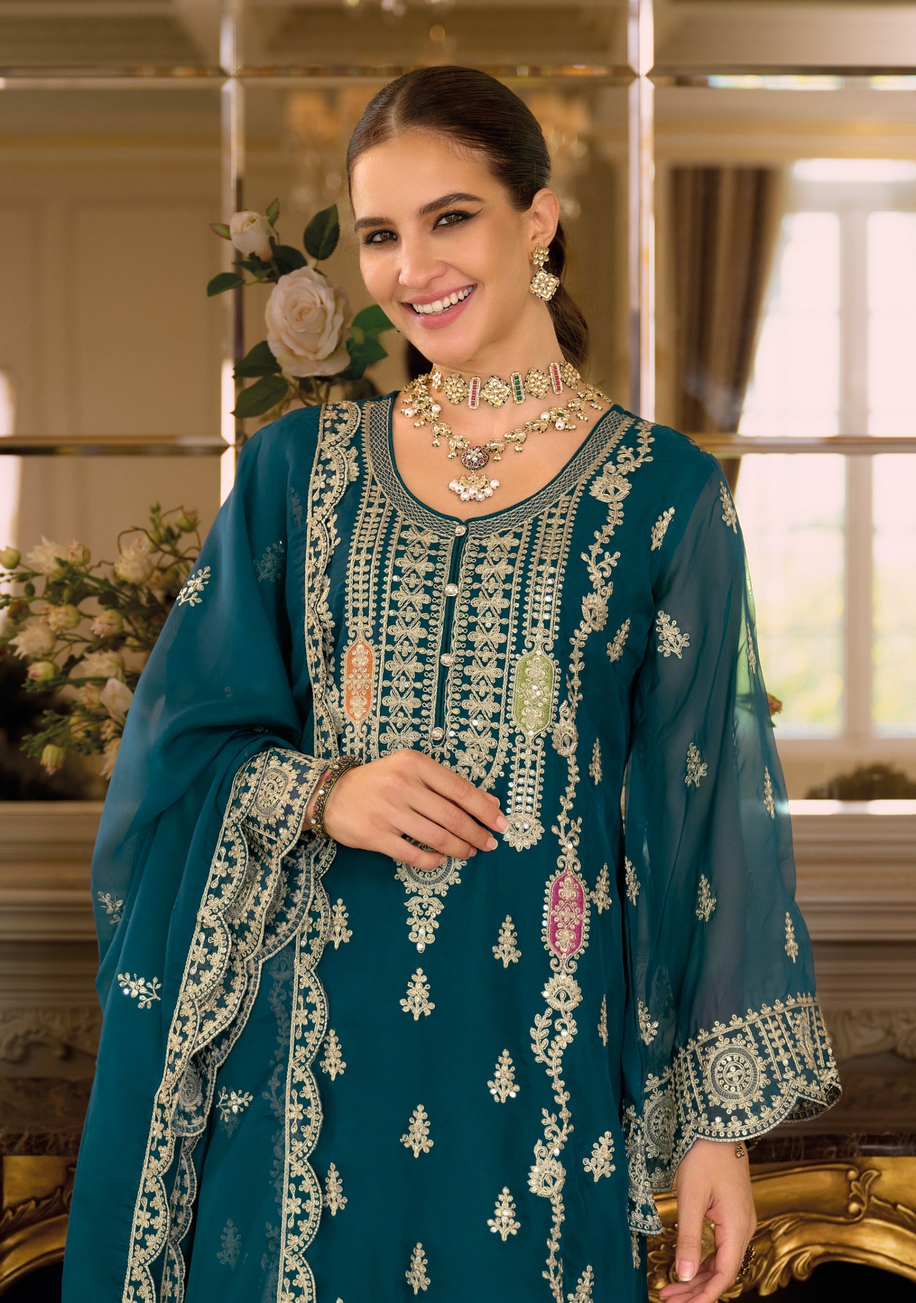 Woman wearing a teal blue embroidered traditional outfit with gold jewelry indoors.