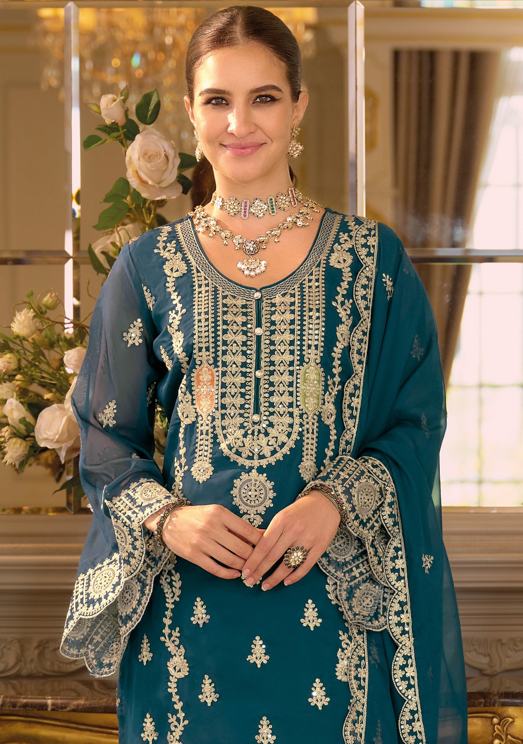 Woman wearing a teal blue embroidered traditional outfit with gold jewelry indoors.