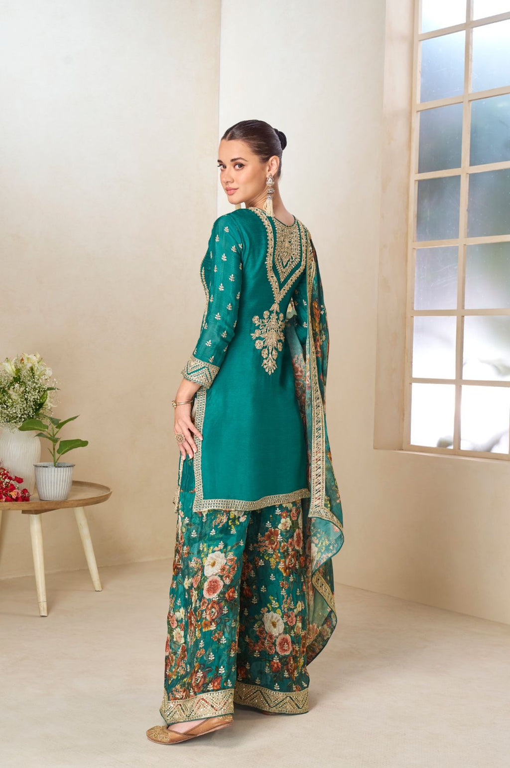 Woman in a traditional teal and floral outfit standing in a room with a window and small table.