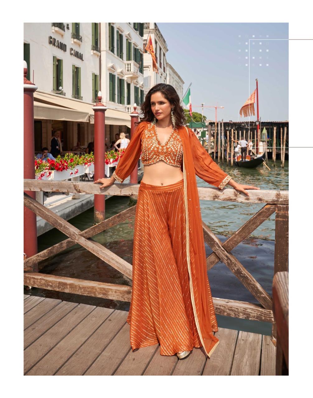 Woman in an orange outfit standing on a wooden pier with a canal and buildings in the background.