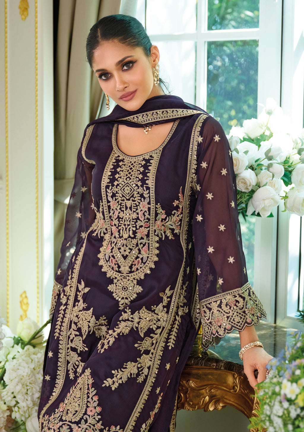 Woman wearing a dark purple embroidered traditional outfit with gold details in a decorated room.