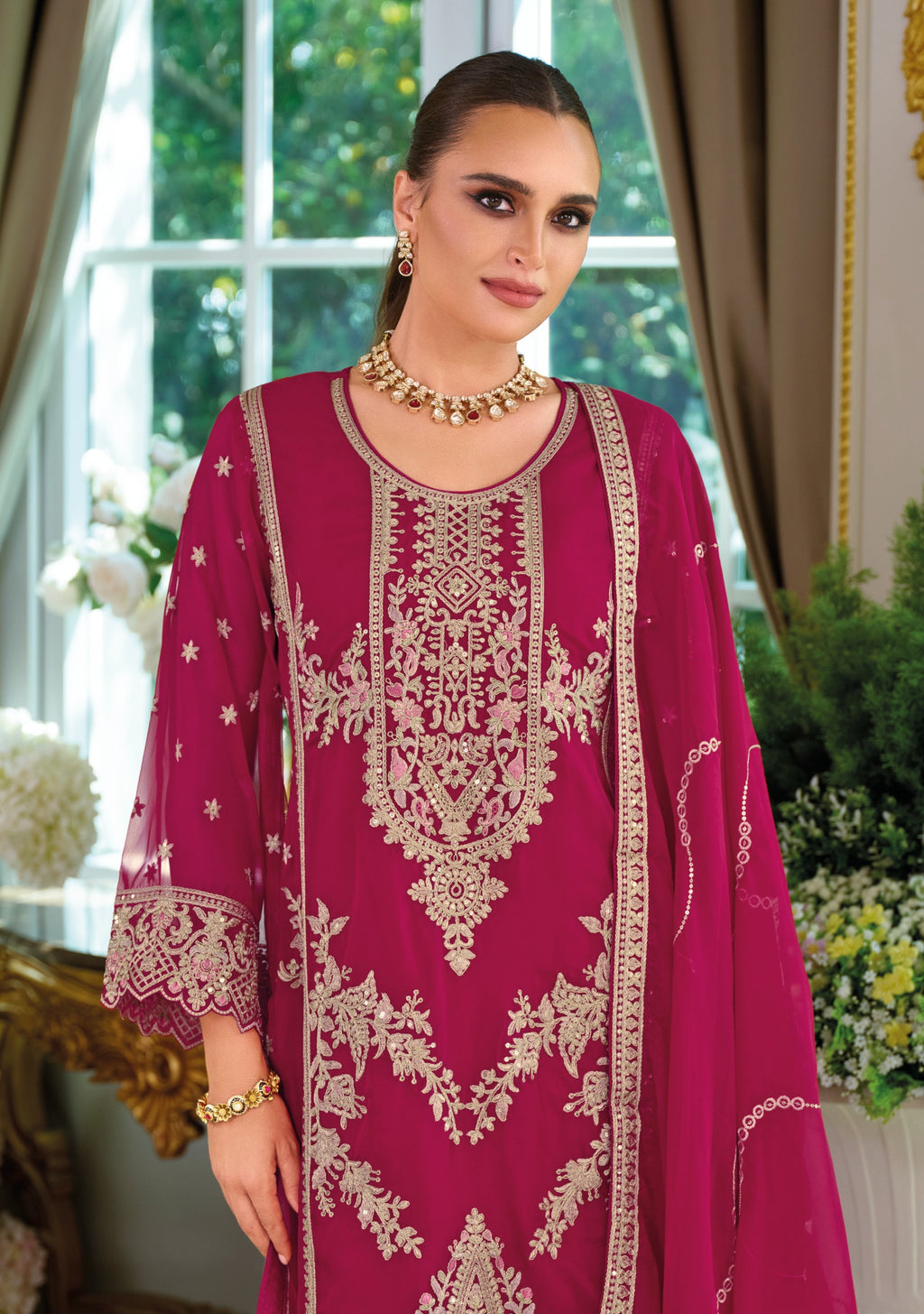 Woman wearing a magenta embroidered traditional outfit in a decorated room.