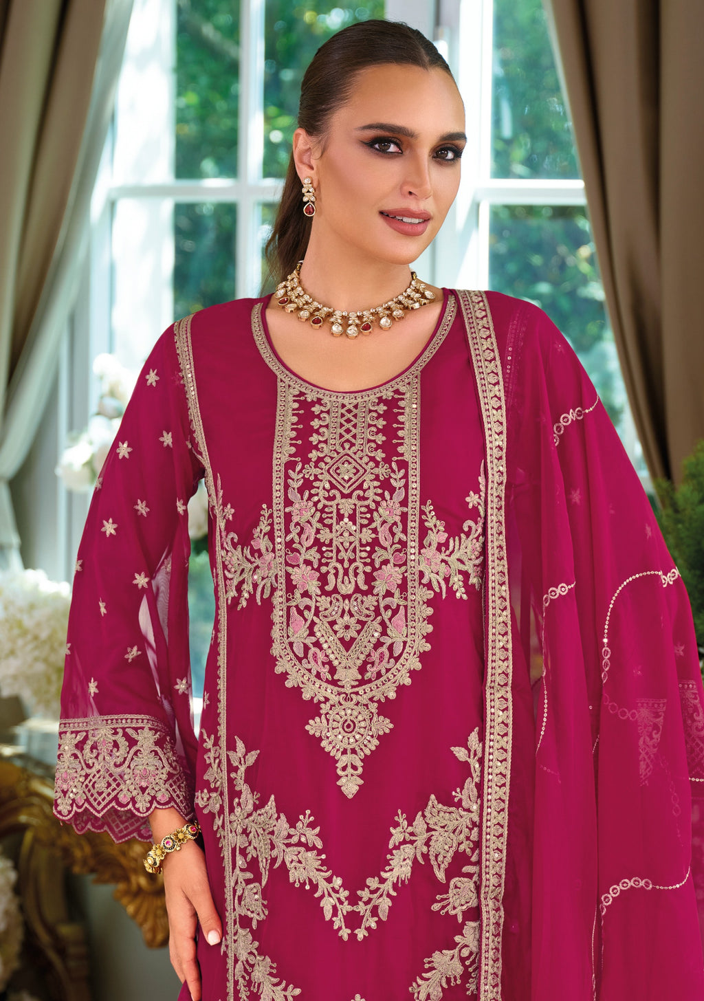 Woman wearing a pink embroidered traditional outfit with gold jewelry indoors.