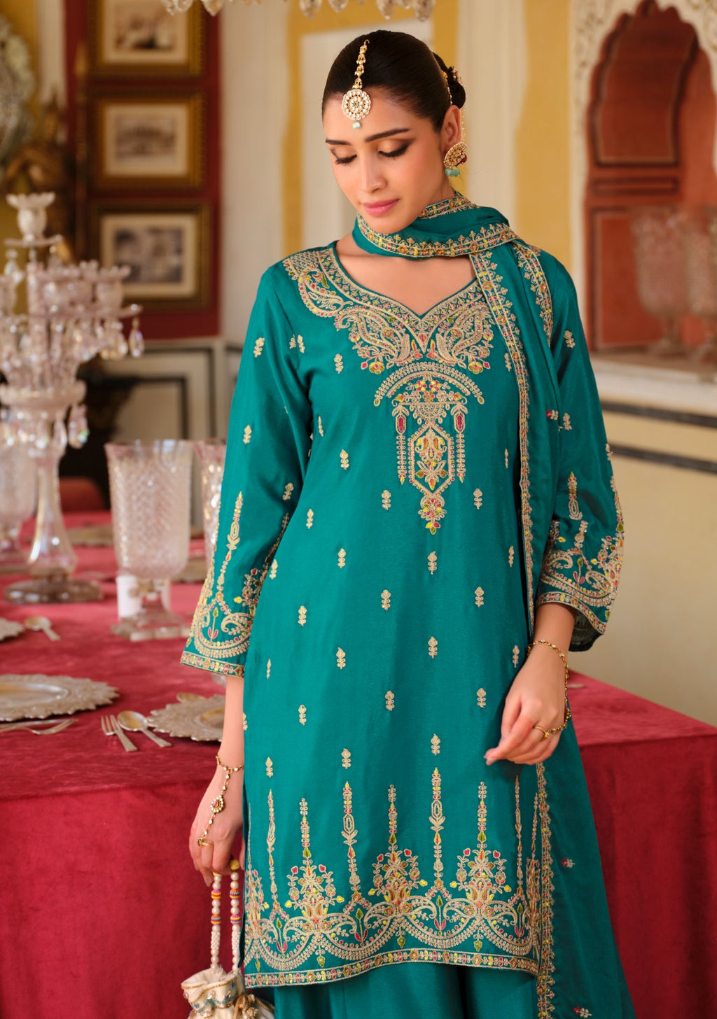 Woman in a teal embroidered outfit standing in an ornate room with red tablecloth.