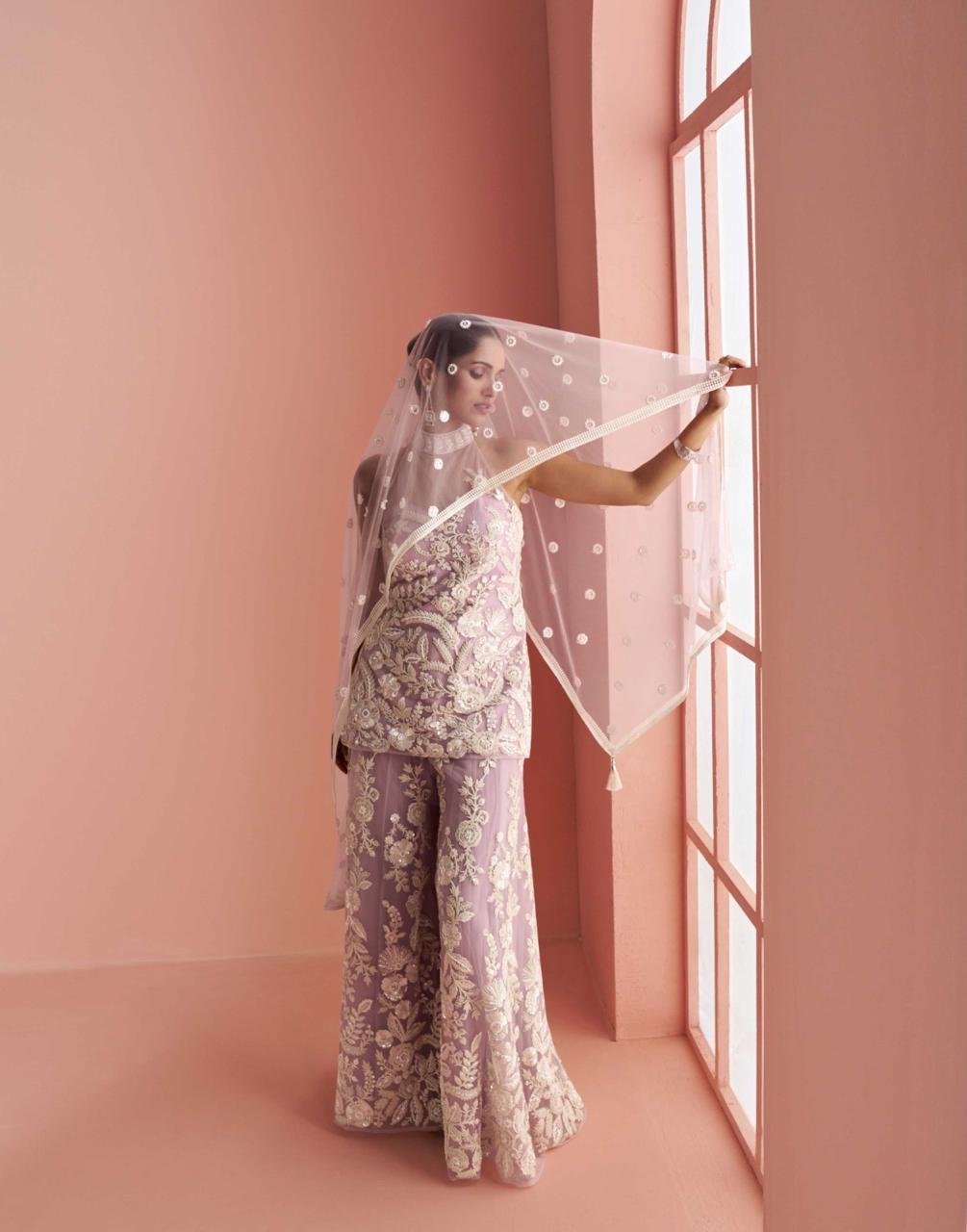 Woman in a floral outfit holding a sheer fabric against a pink wall.