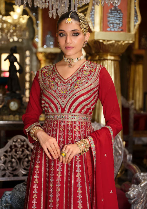 Woman in a red and gold traditional outfit with intricate patterns, standing in an ornate interior setting.